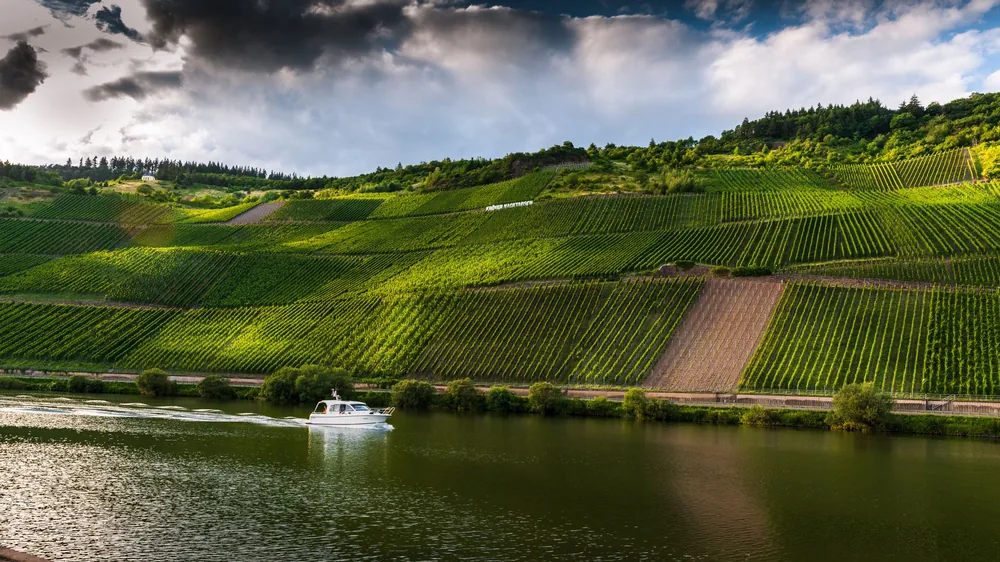 A boat glides along a river beside lush vineyards under dramatic clouds