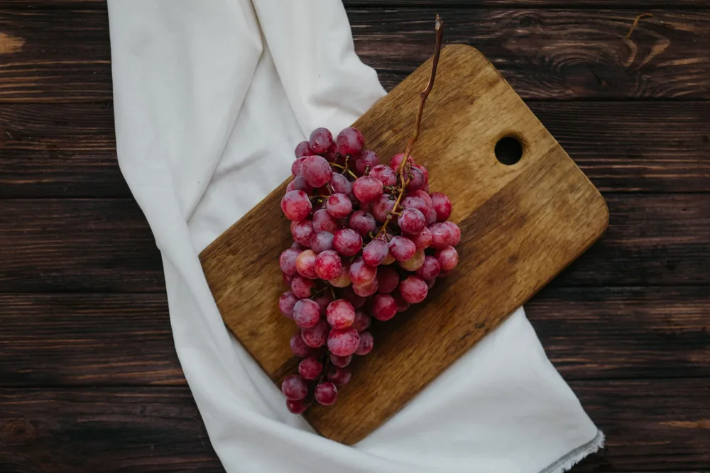 A bunch of fresh red grapes placed on a rustic wooden cutting board with a cloth background