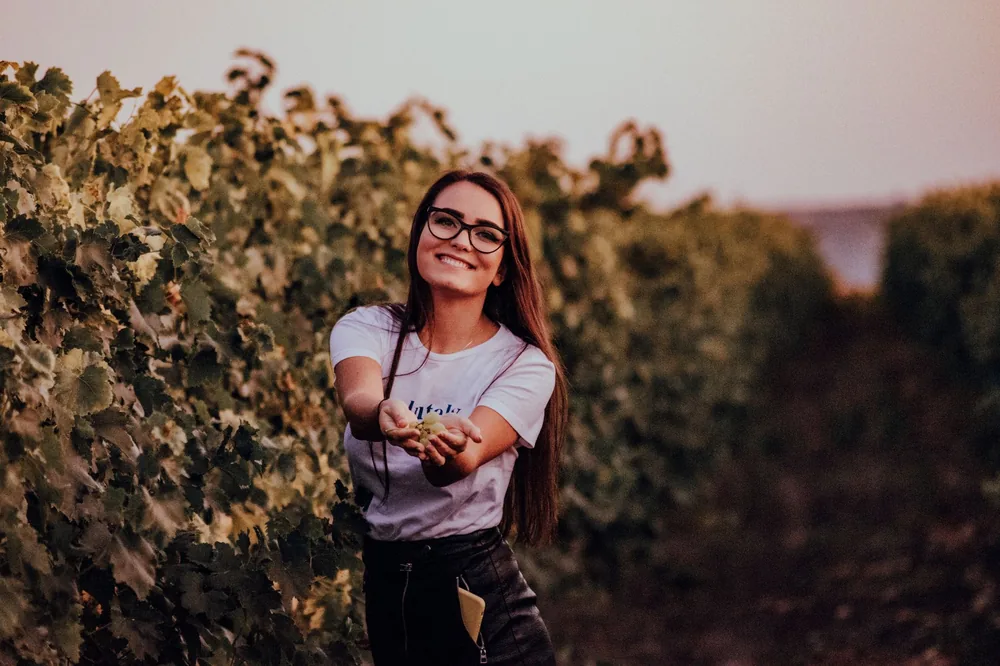 A cheerful woman standing in a vineyard, holding grapes and smiling warmly