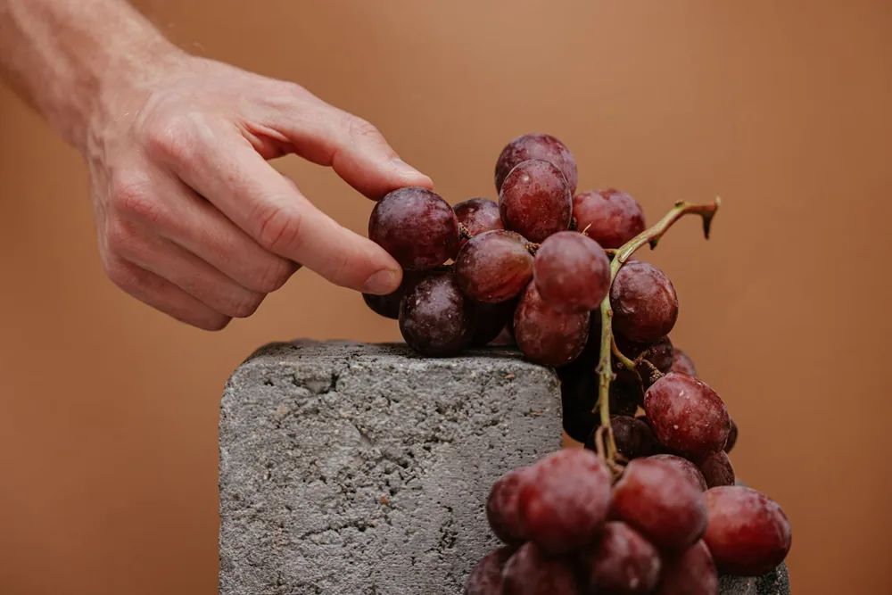 A close-up shot of red grapes and a hand on a stone block against a brown background
