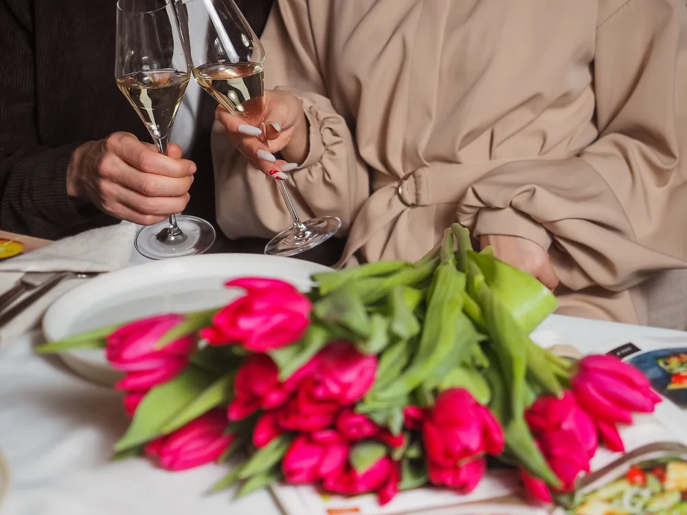 A couple toasting with champagne glasses over a bouquet of pink tulips on a dining table