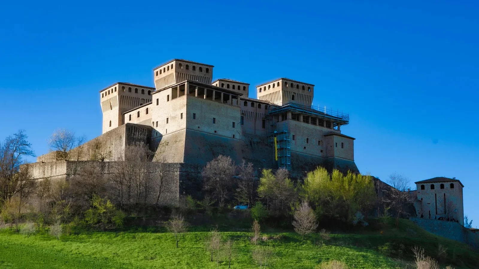A grand medieval castle sits atop a hill against a clear blue sky, surrounded by lush greenery