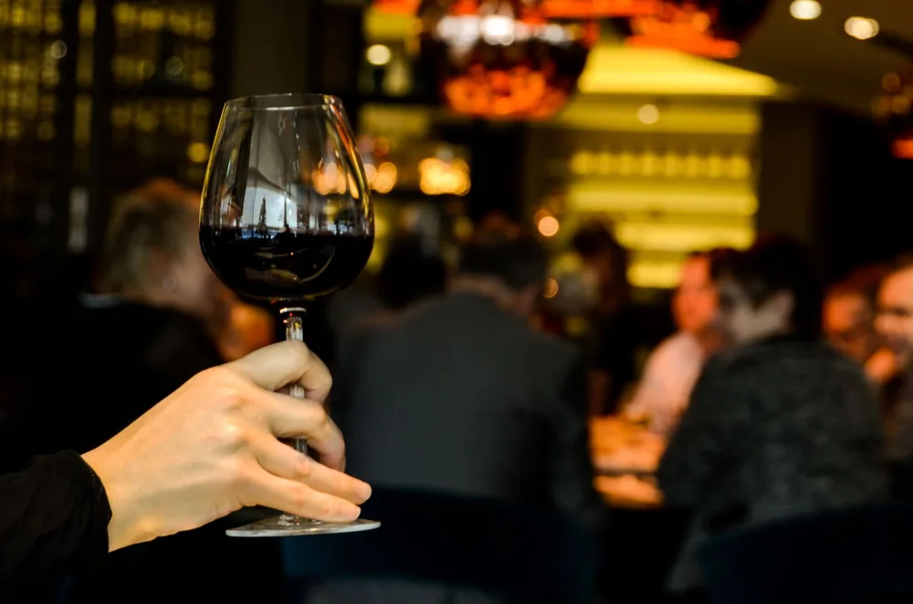 A hand holding a glass of red wine with a blurred group dining in a stylish restaurant