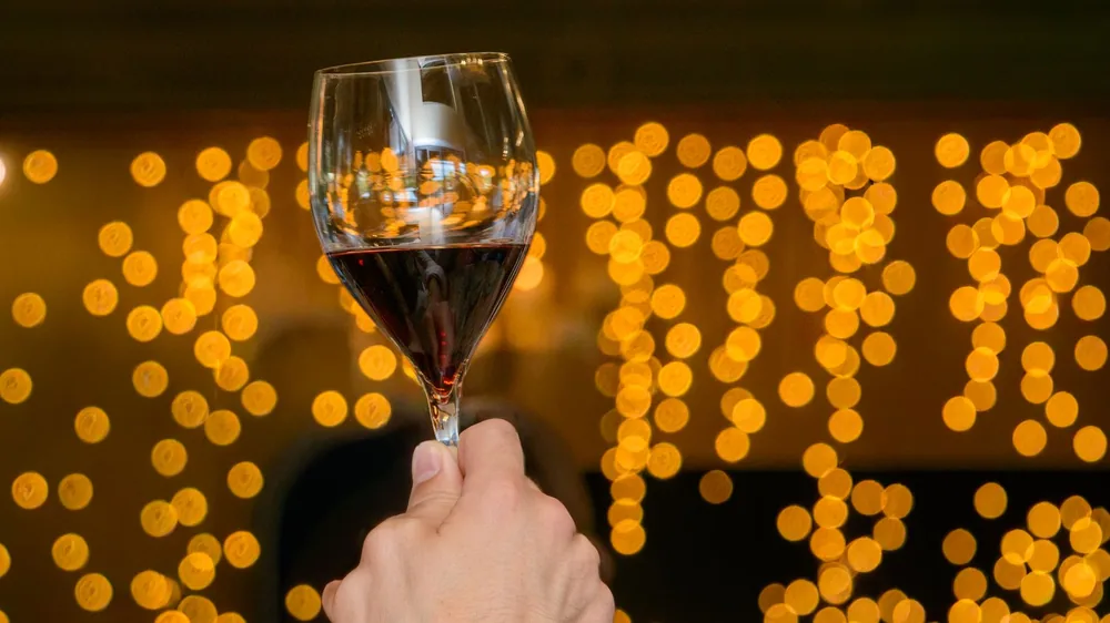 A hand holding a red wine glass against warm bokeh lighting, creating a festive atmosphere