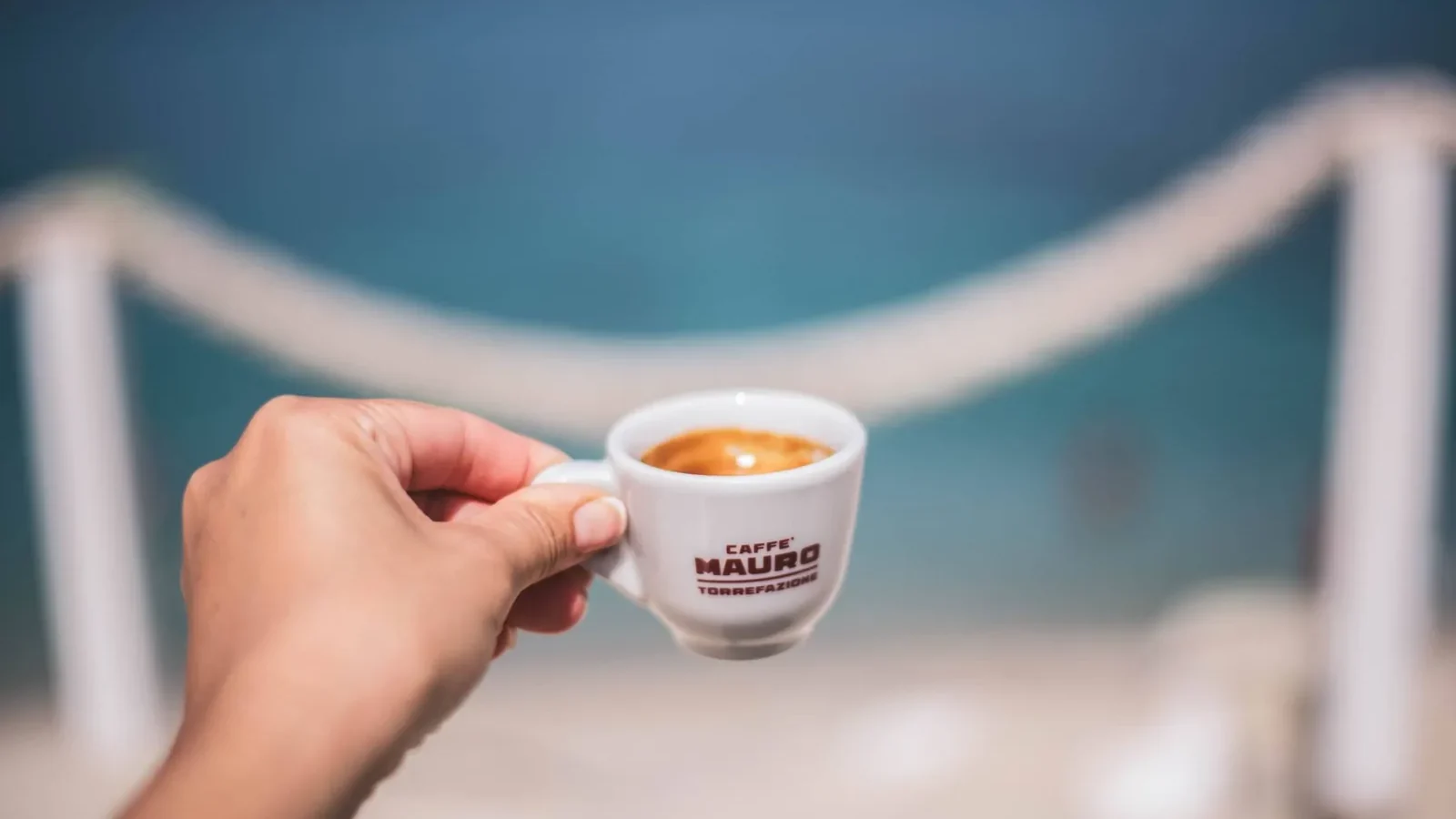 A hand holding an espresso cup over a blurred ocean background, capturing a serene coastal moment