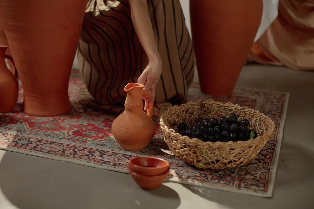 A hand reaches for a clay jug next to grapes in a wicker basket on an ornate carpet