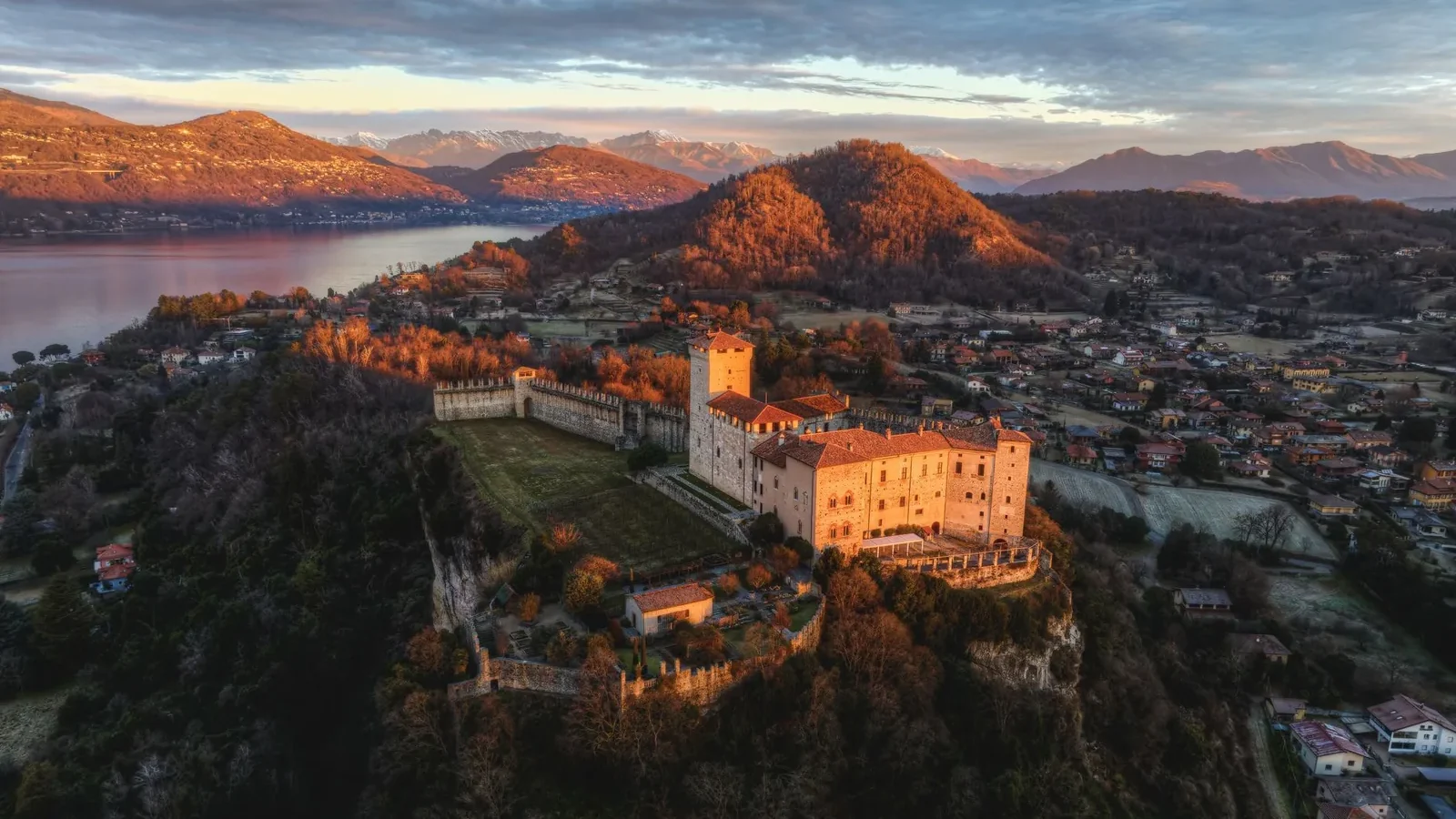 A historic castle overlooking a lake and village at sunset, surrounded by mountains