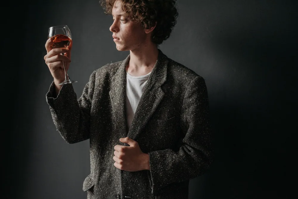 A man holding a wine glass, wearing a gray blazer, expressing focused observation