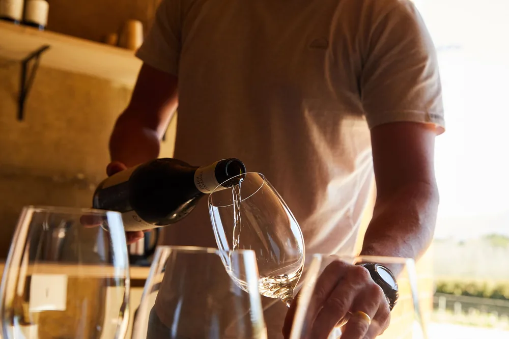 A man pours white wine into glasses indoors during sunset, creating a warm ambiance