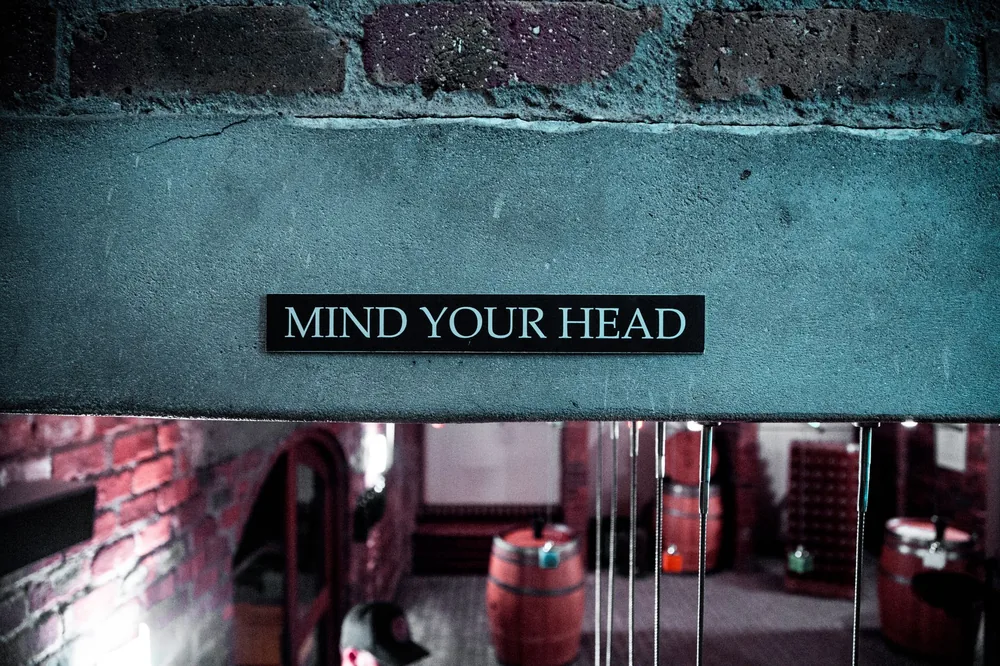 A 'Mind Your Head' sign in a rustic brick barrel cellar setting, creating a safety awareness ambiance