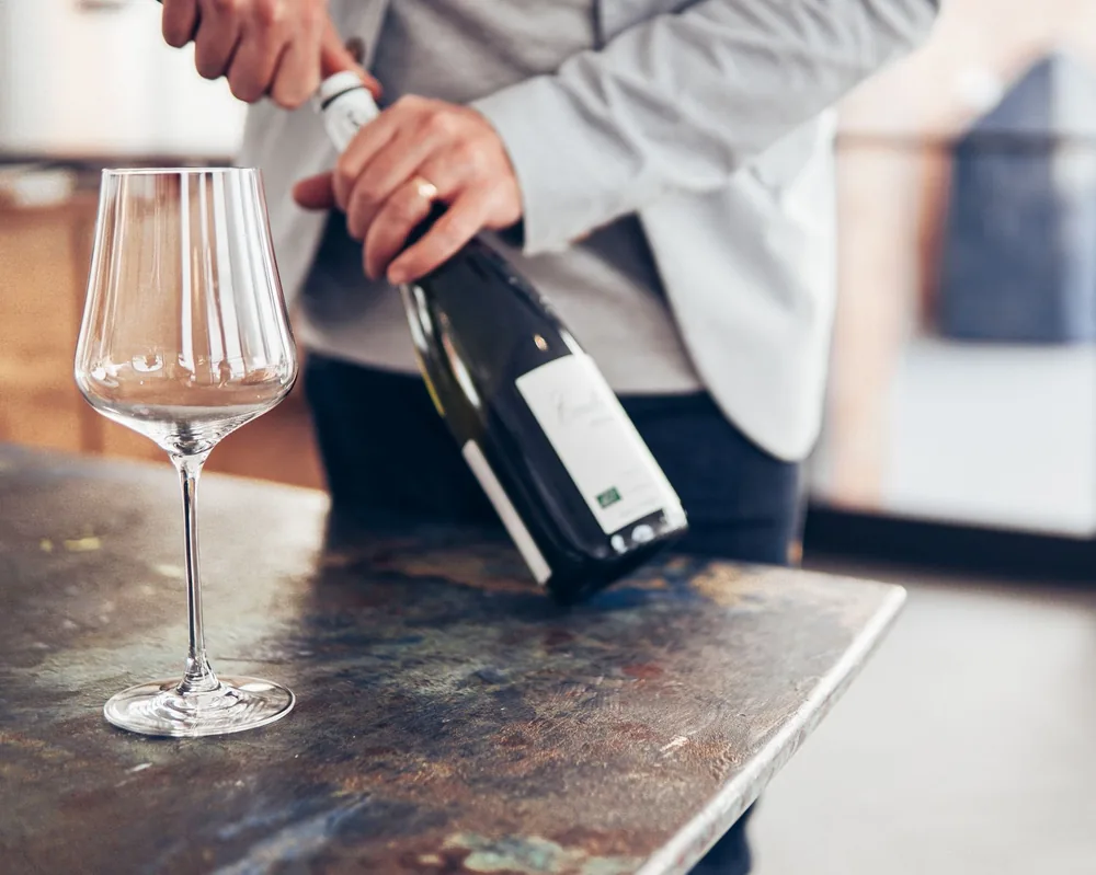 A person elegantly opening a wine bottle beside an empty glass on a modern table