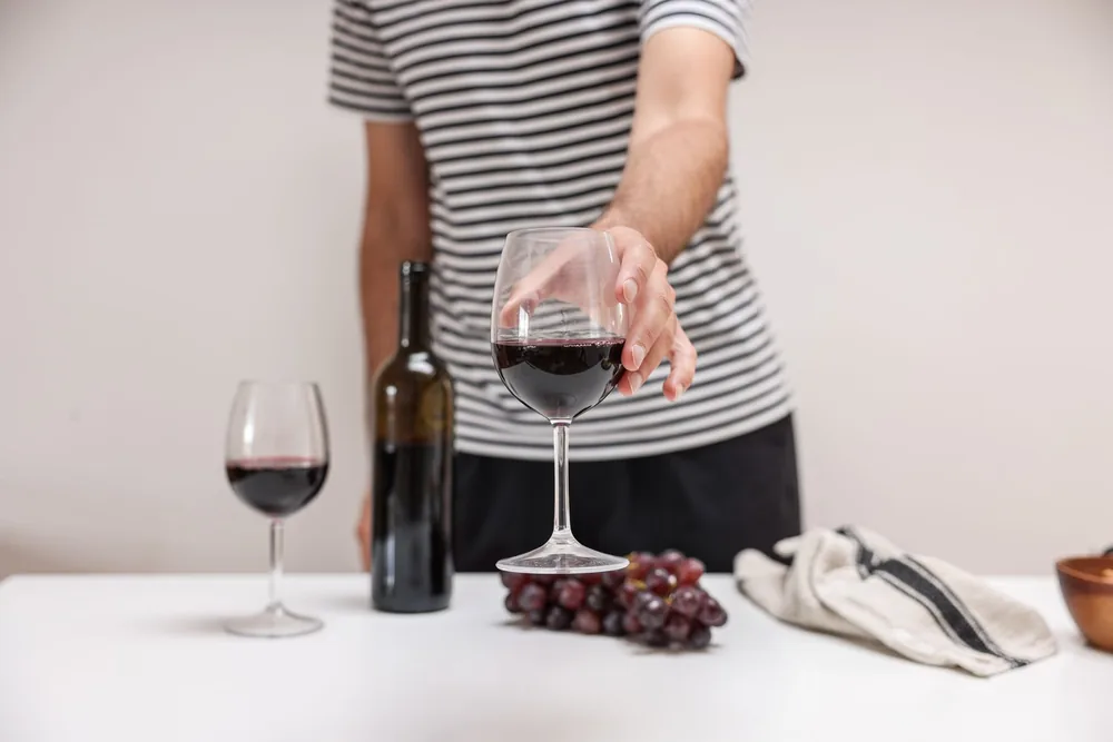 A person in a striped shirt holding a glass of red wine next to grapes and a wine bottle