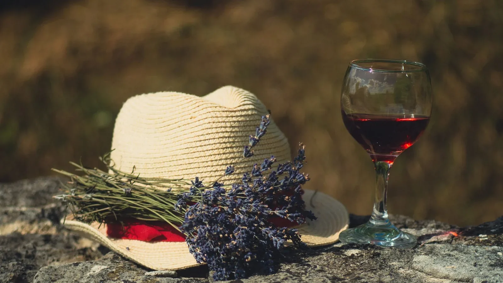 A relaxing outdoor scene with red wine, lavender, and a straw hat on a stone surface