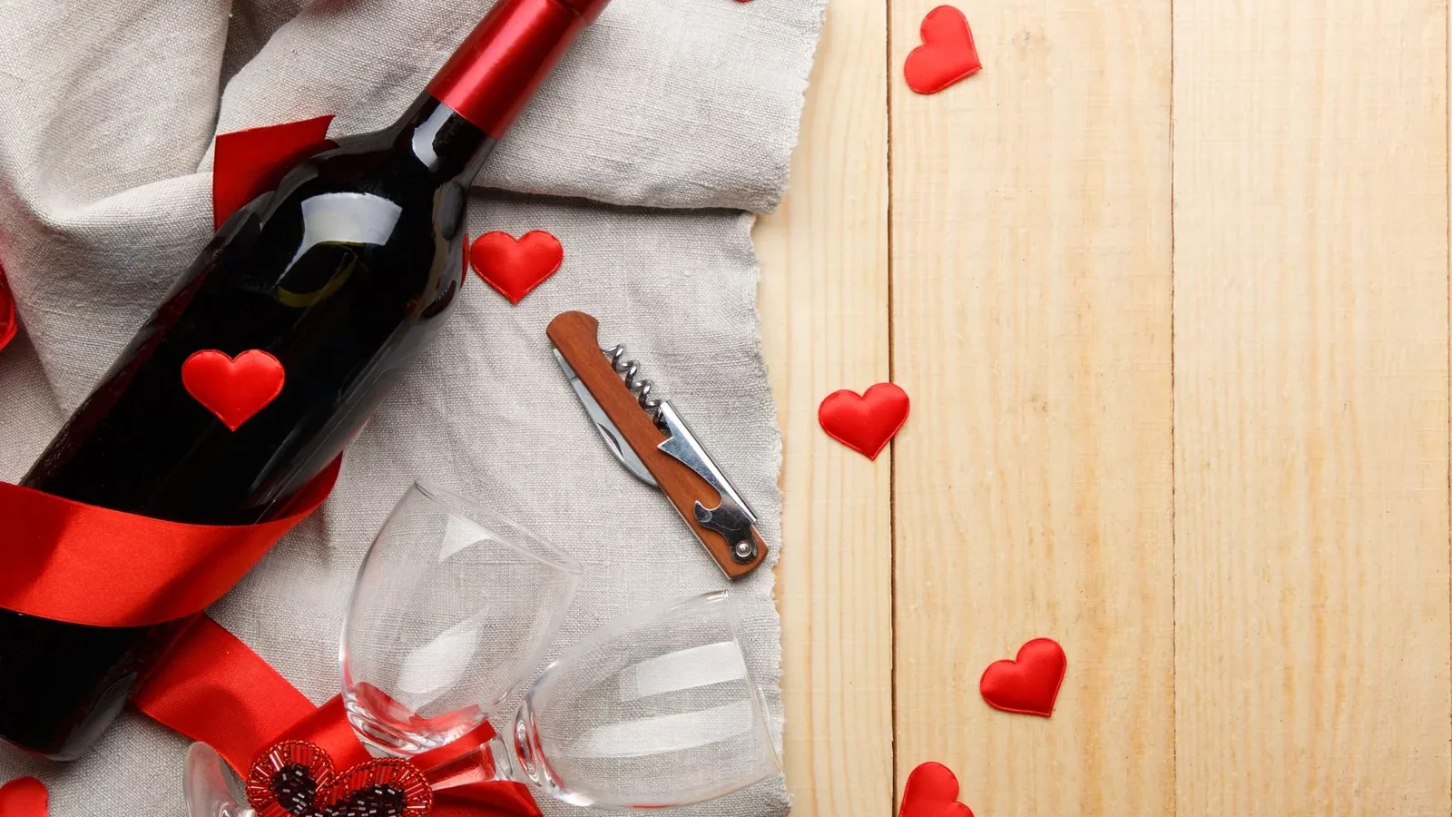 A romantic flat lay of wine bottle, glasses, and red hearts on a wooden surface with burlap
