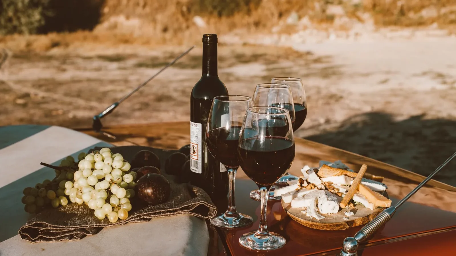 A rustic outdoor picnic setup featuring wine, grapes, and cheese on a car bonnet