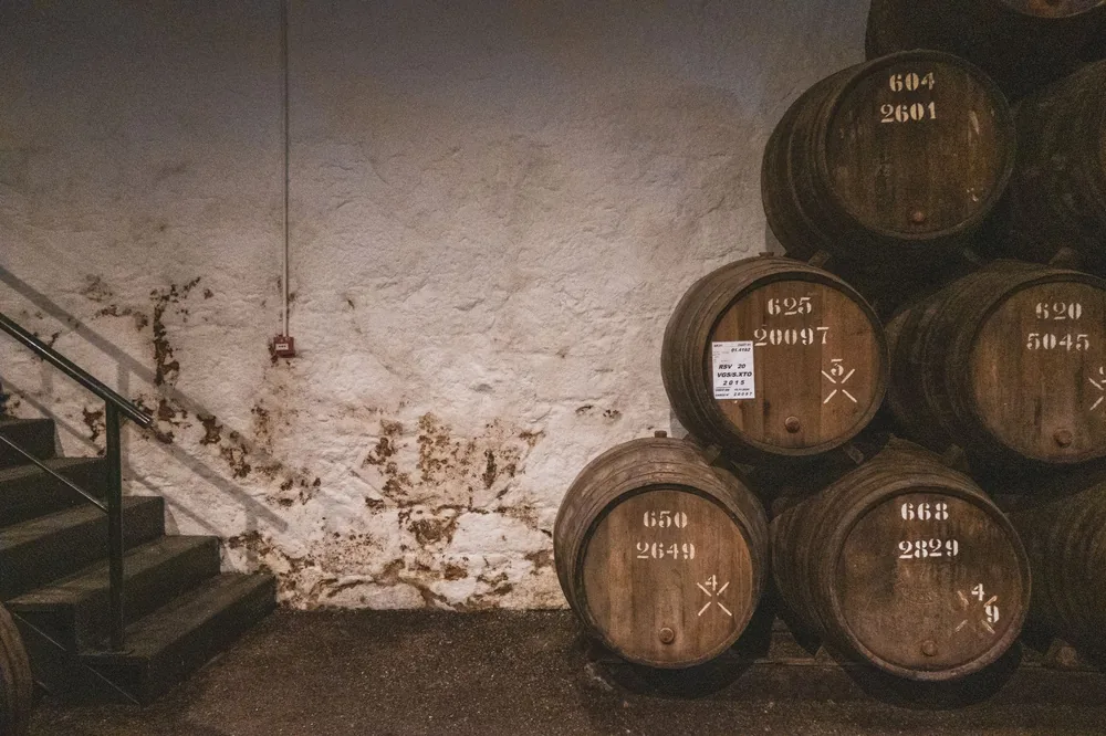 A stack of aged wooden barrels in a rustic distillery cellar with stone walls and stairs