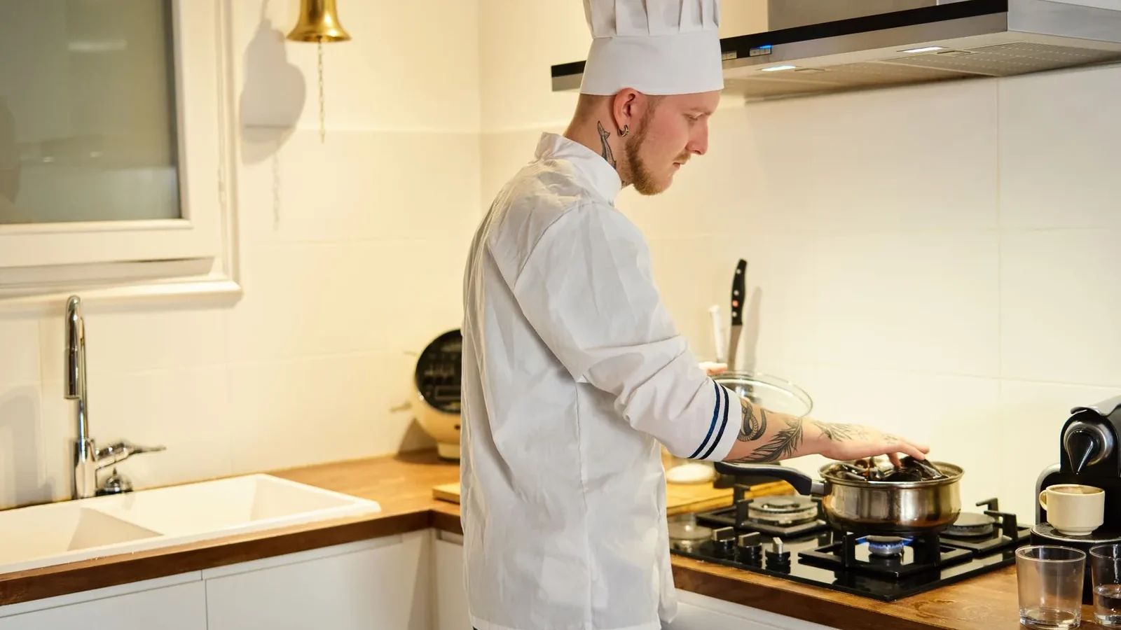 A tattooed chef wearing a hat cooks in a modern kitchen, focusing on a pot on the stove