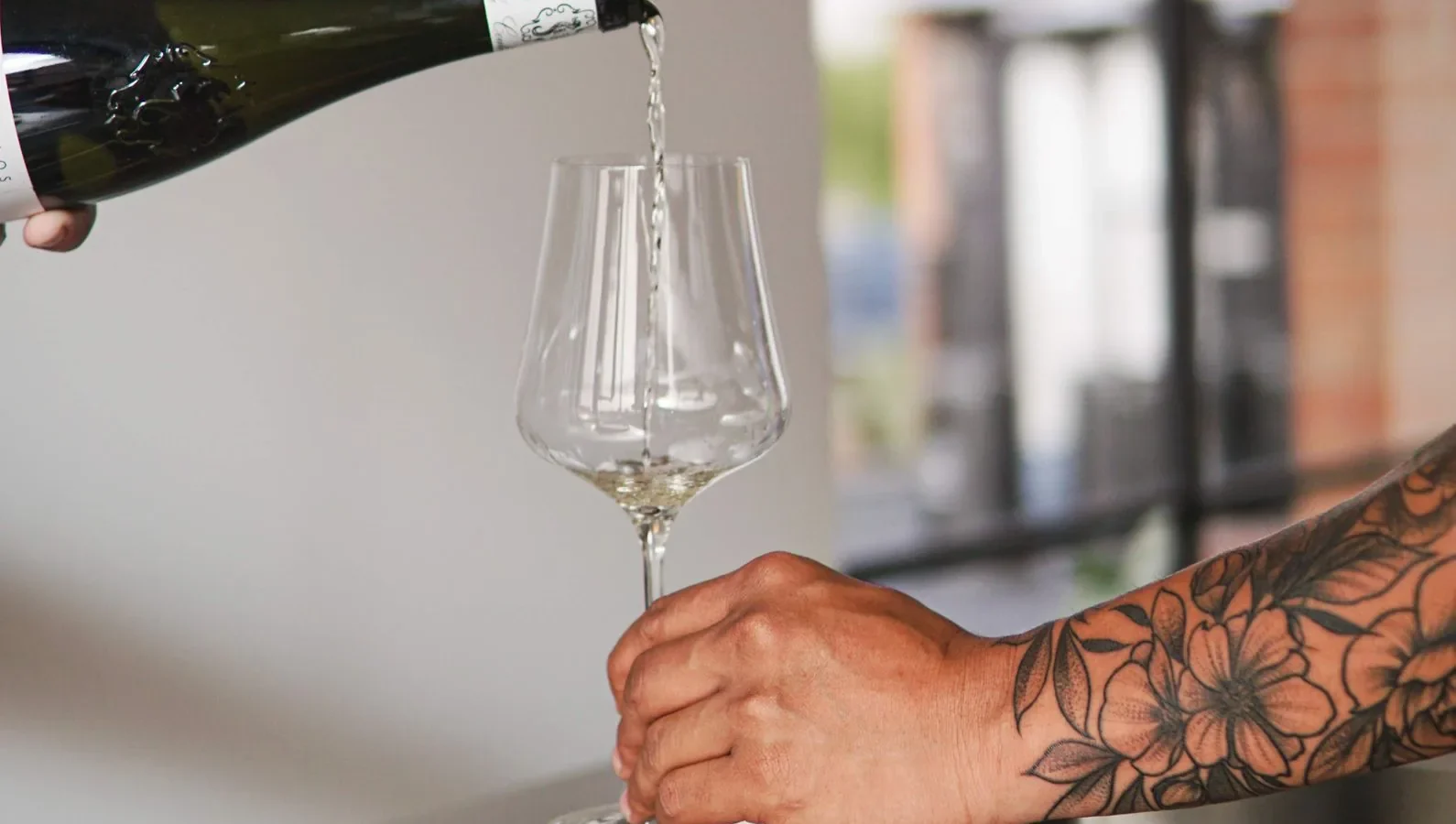 A tattooed hand holding a wine glass while champagne is poured indoors, creating a festive ambiance