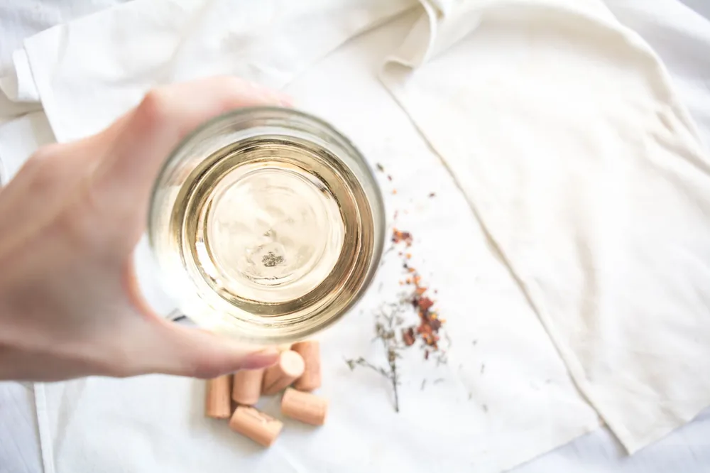 A top-down shot of a hand holding a glass of white wine with corks and a white cloth in the background