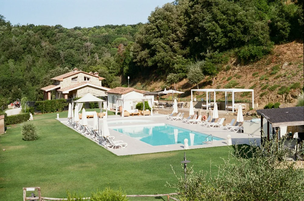 A Tuscan farmhouse and its pool sit cradled by green hills under a clear summer sky