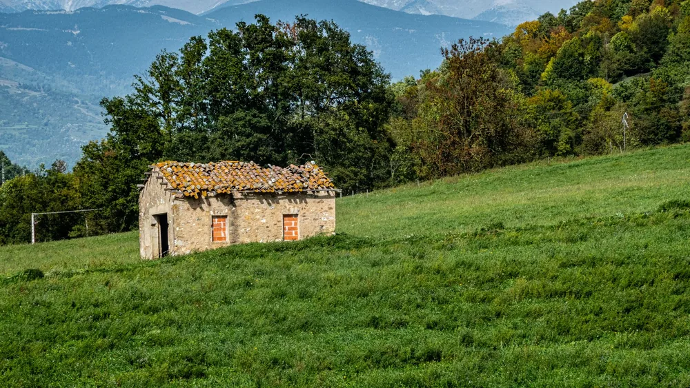 A vintage stone cottage surrounded by lush greenery and mountains under a clear blue sky