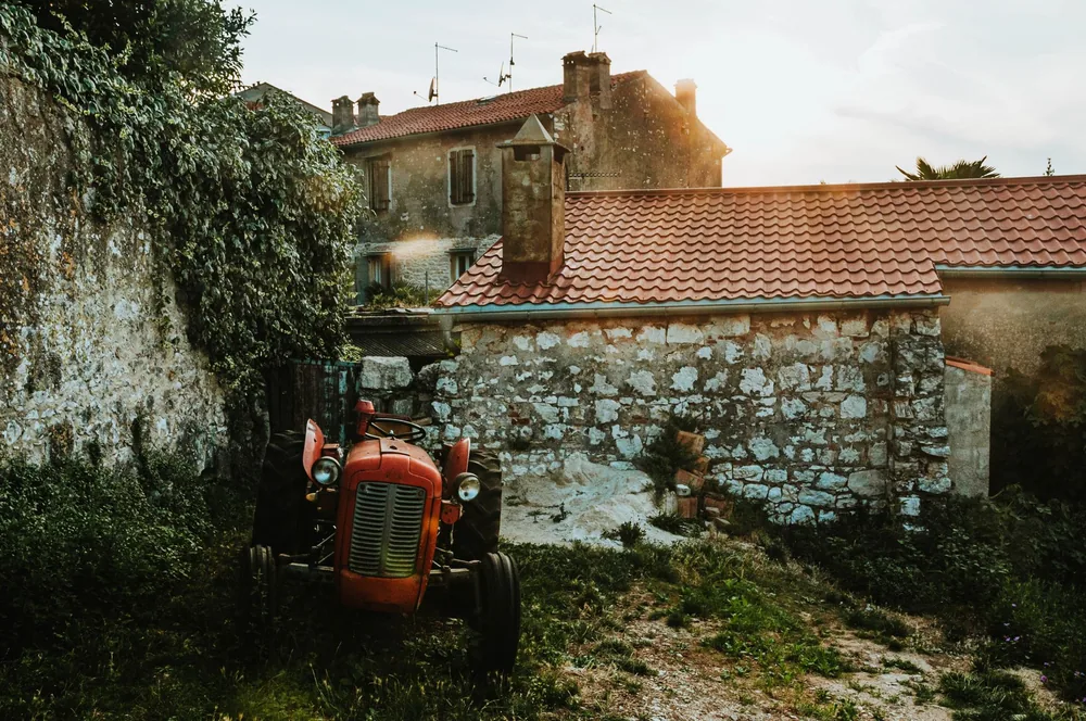 A vintage tractor sits near a rustic stone farmhouse, captured during sunset, conveying a serene countryside feel