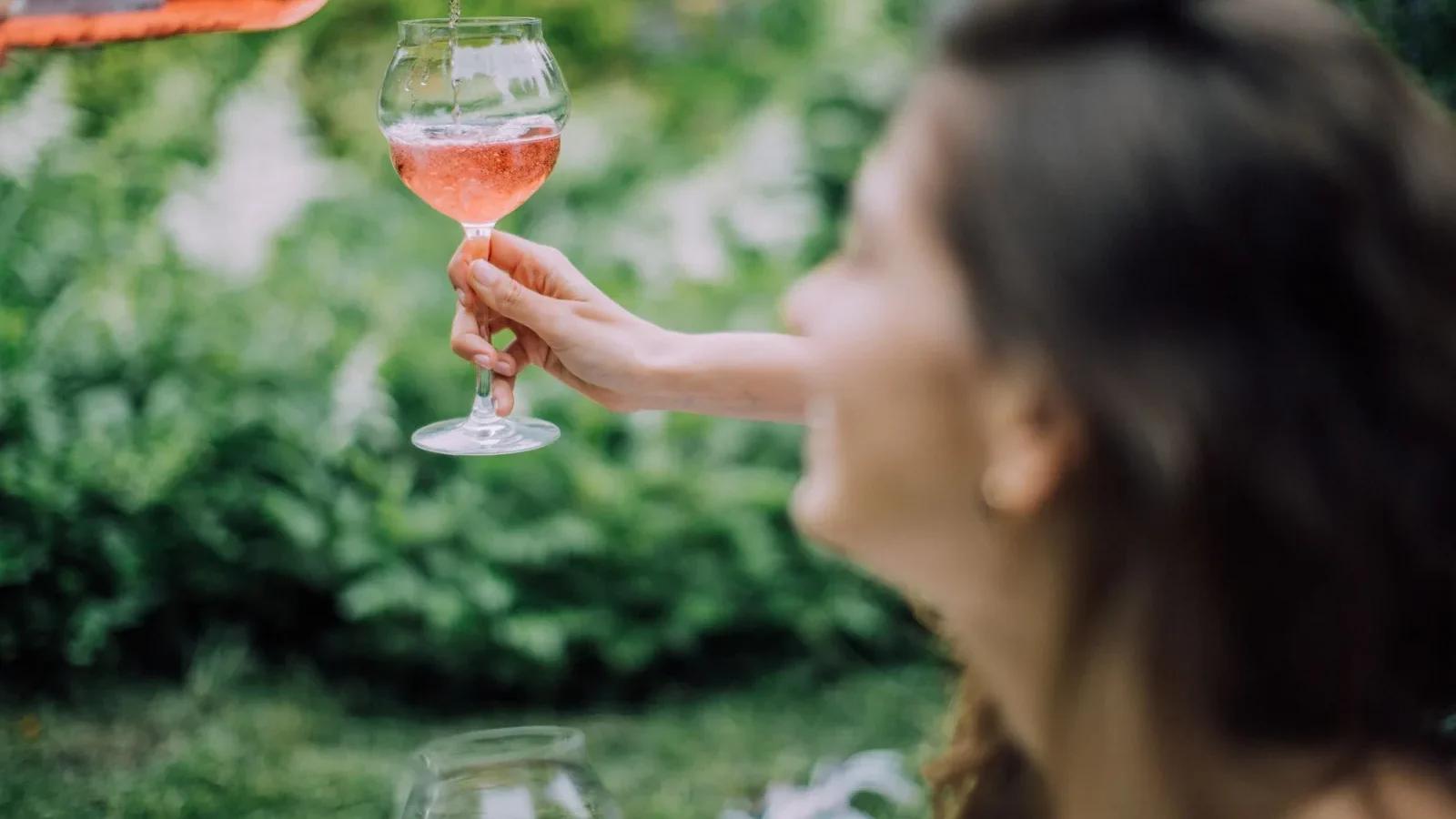 A woman elegantly pours rosé wine into a glass during a sunny outdoor picnic, capturing a relaxed summer vibe
