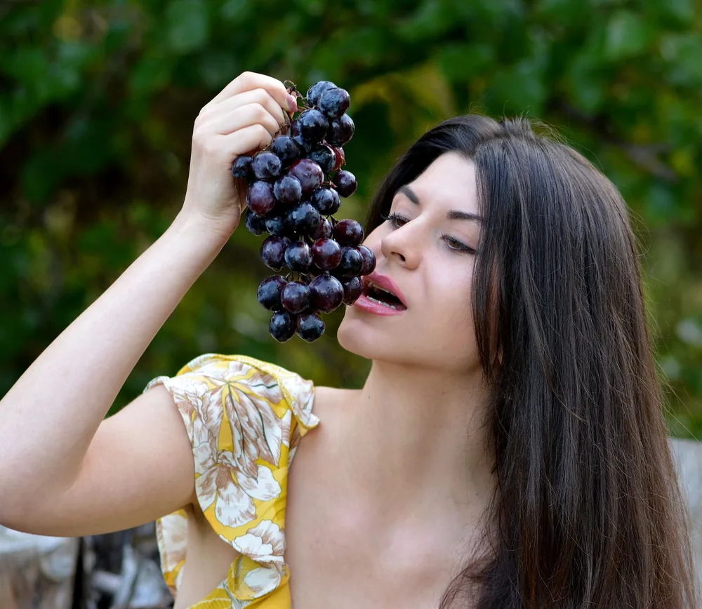 A woman in a floral dress enjoying fresh black grapes outdoors, showcasing natural beauty and healthy living