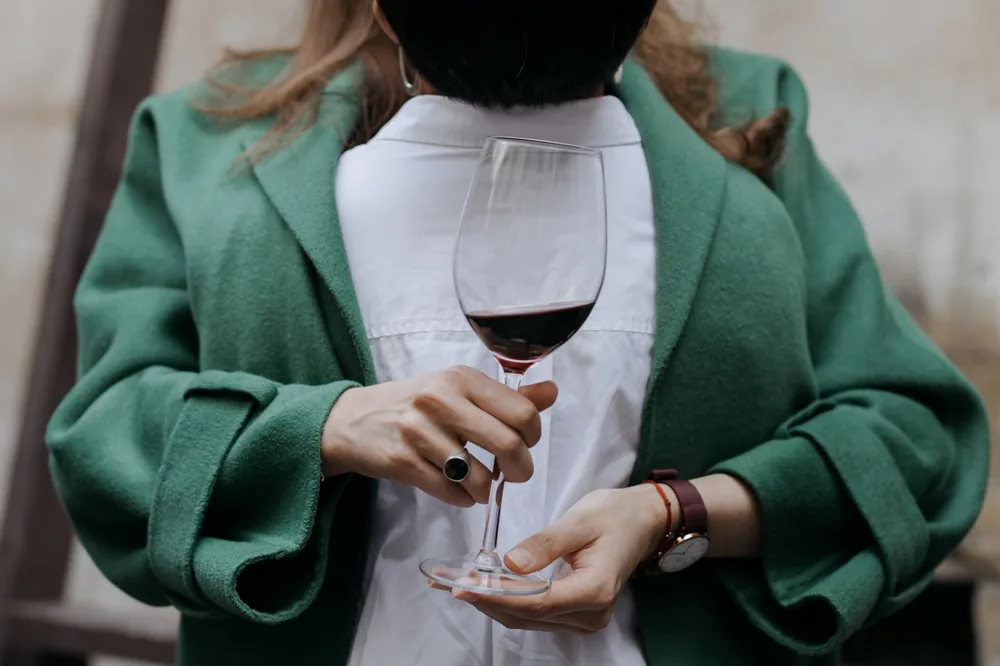 A woman in a green coat holds a wine glass filled with red wine, showcasing elegance and style