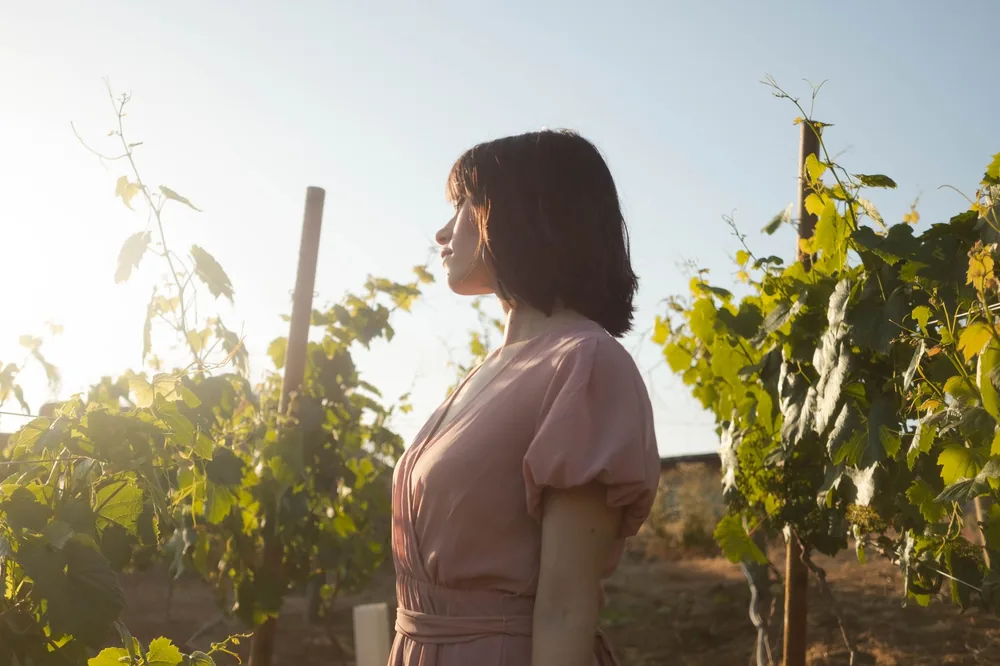 A woman in a pink dress stands in a vineyard, basking in the warm sunset light