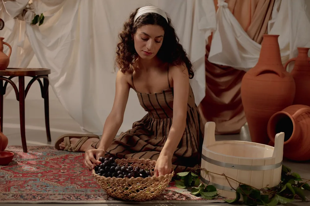 A woman in a rustic setting sorting grapes, surrounded by ceramics and drapery. Indoor studio shot