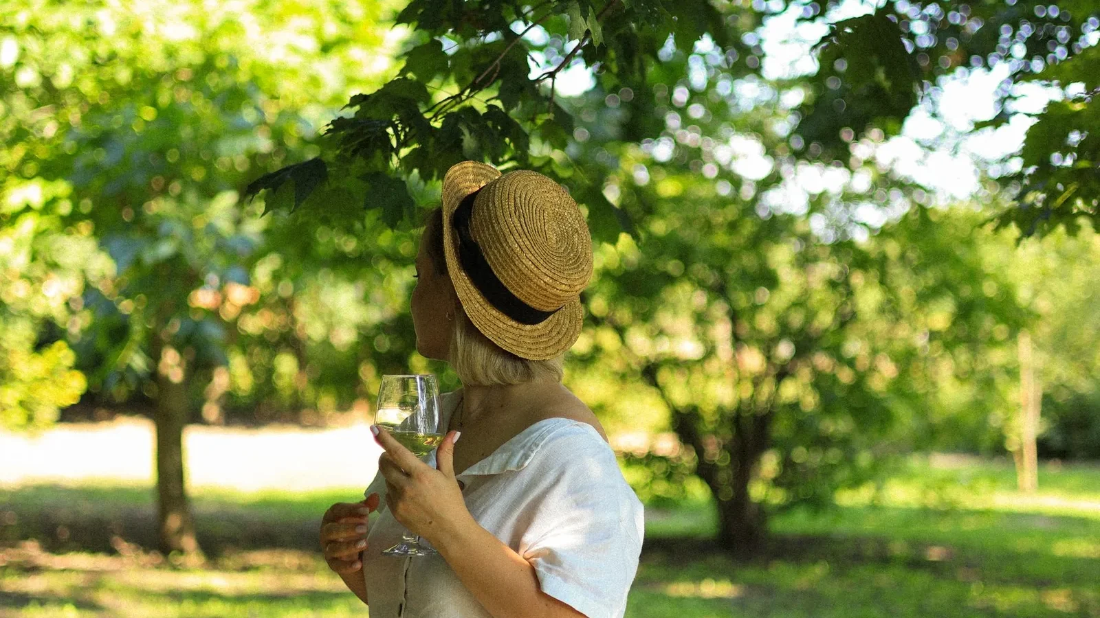 A woman in a straw hat holding a wineglass, enjoying a sunny day in a green park