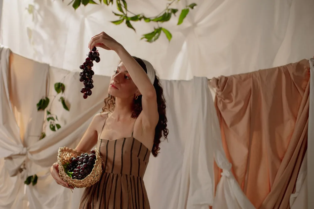 A woman in a striped dress holds grapes in a rustic, draped setting