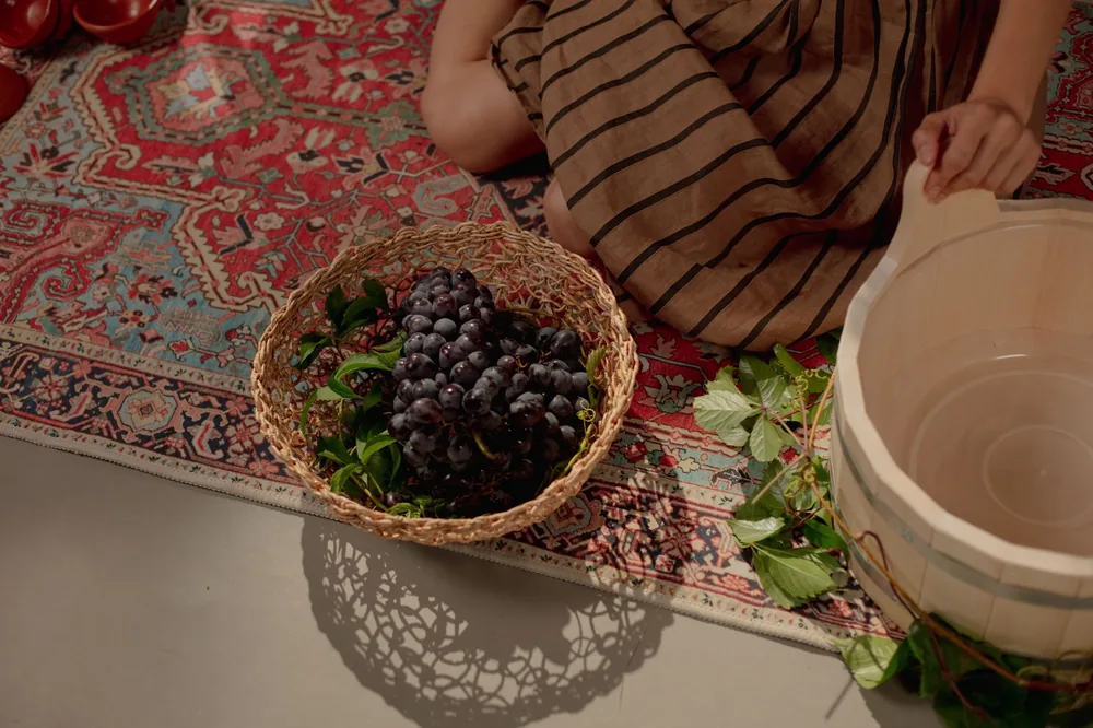 A woman in a striped dress touches grapes in a basket beside a wooden vat on a colorful carpet