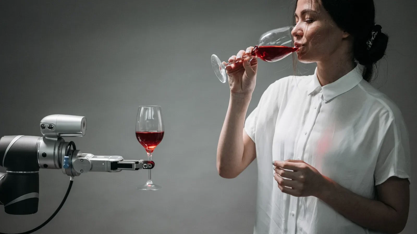 A woman in a white shirt drinking red wine interacts with a robot arm holding a wine glass