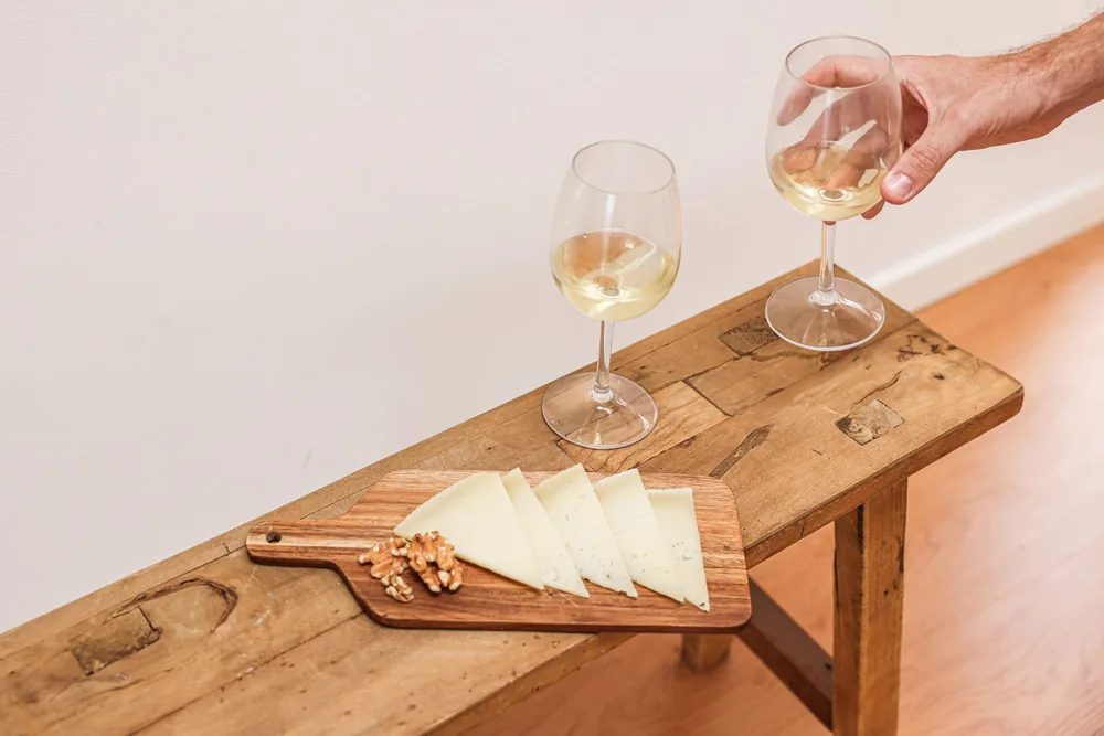 A wooden board with sliced cheese, walnuts, and white wine glasses on a rustic table