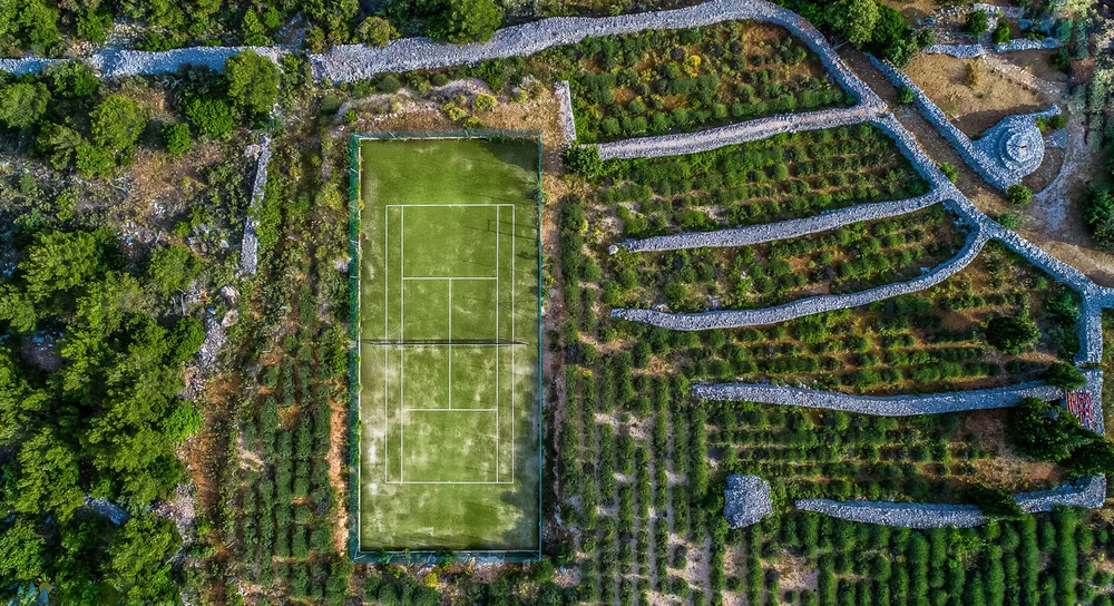Aerial view showcasing a tennis court surrounded by a terraced landscape with greenery and stone walls, ideal for outdoor sports