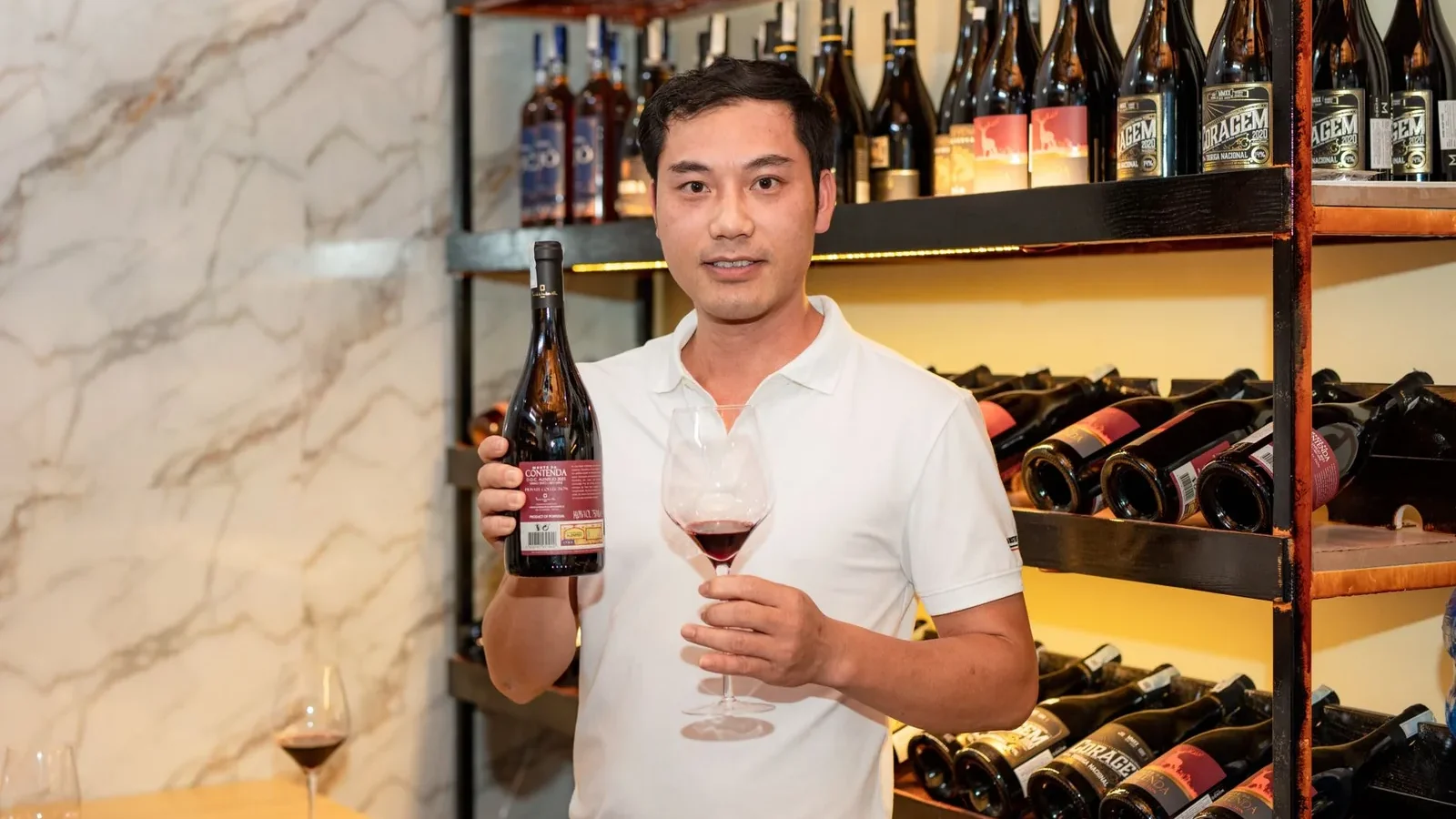 An adult man in a liquor store holding a wine bottle and glass, surrounded by wine racks