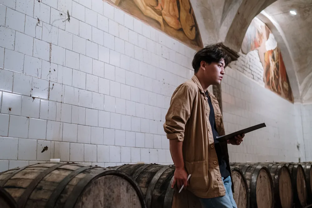 An expert inspecting wooden fermentation barrels in a brewery indoors, with a checklist in hand