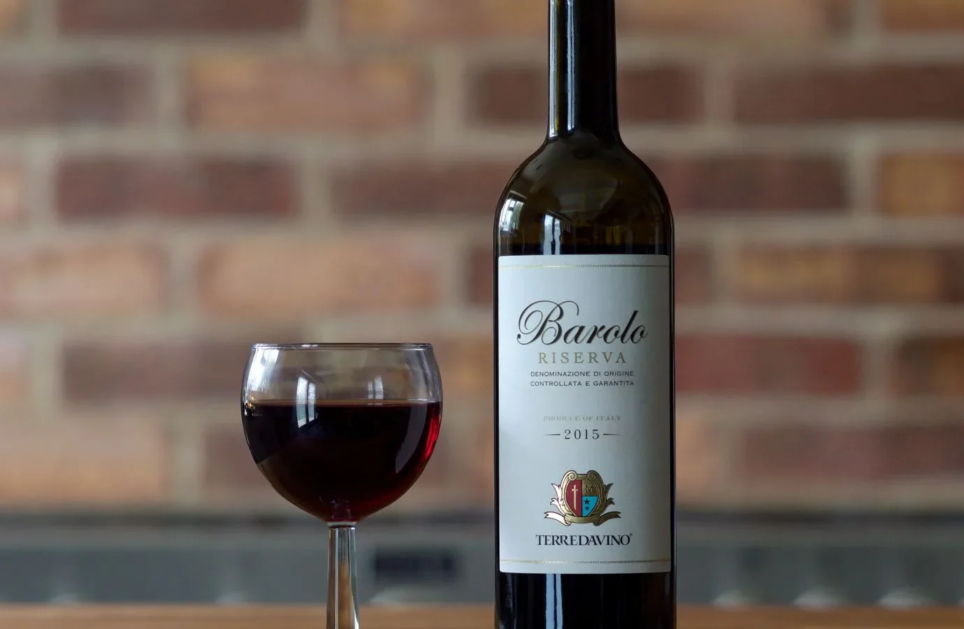 Barolo Riserva 2015 bottle with wine glass on a wooden table against a brick wall