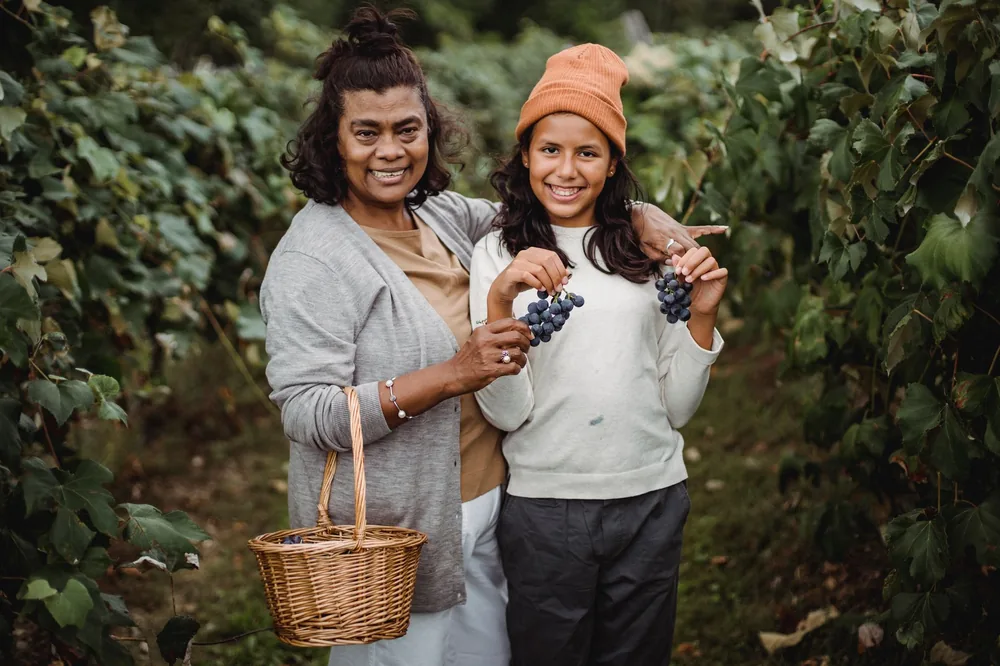 Cheerful ethnic mother with wicker basket embracing charming girl with bunches of fresh grapes between vines while looking at camera