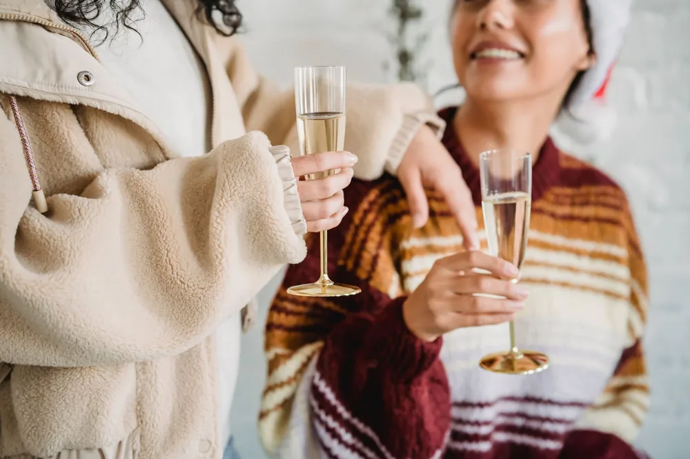 Crop smiling young females in soft sweaters and Santa hat raising glasses of champagne and toasting while celebrating Christmas together