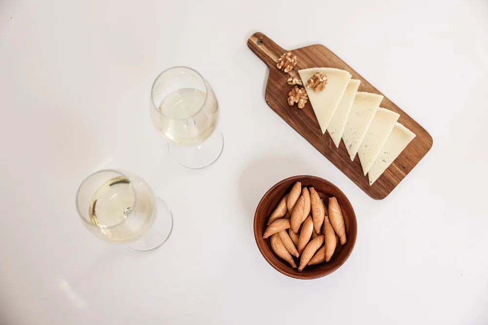 Flat lay of wine glasses, sliced cheese, breadsticks, and walnuts on a wooden board