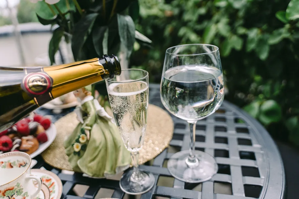 From above of unrecognizable person pouring sparkling wine into elegant crystal glass served on round table near fresh pasties with berries and coffee cups