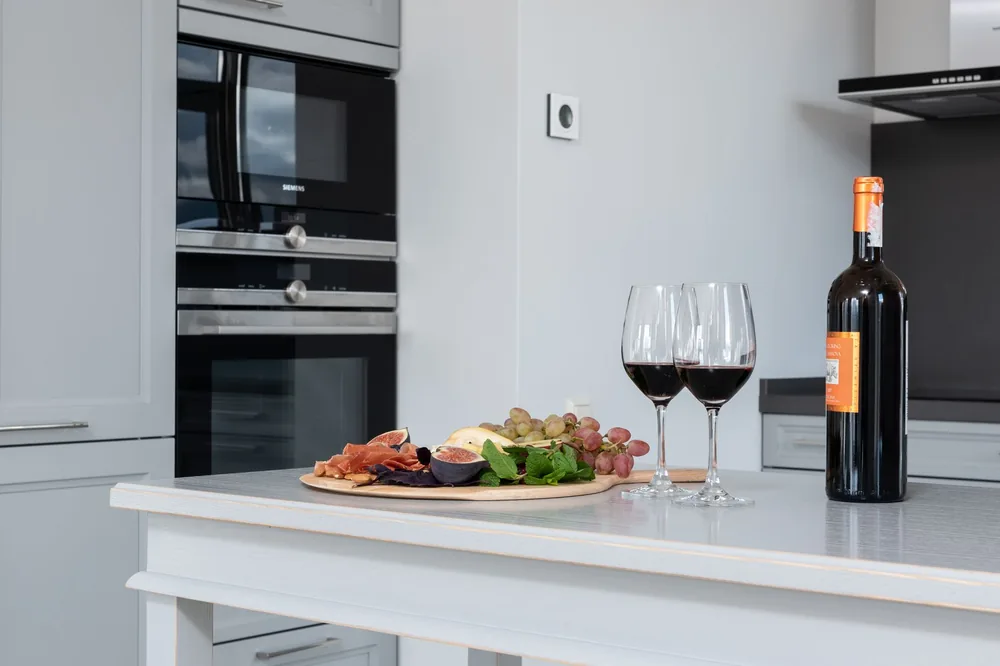 Goblets of red wine placed on white counter with bottle of alcohol beverage and tray of assorted appetizers in kitchen with modern appliances