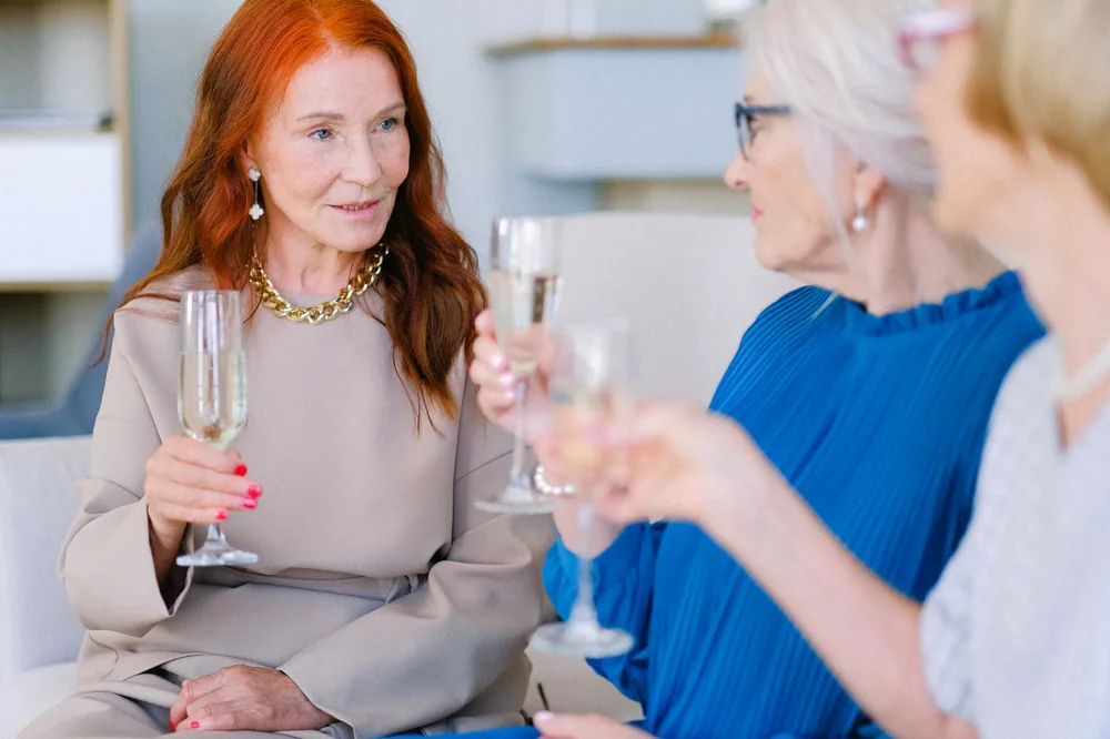 Group of aged crop women making toast with glasses of champagne on blurred background of living room