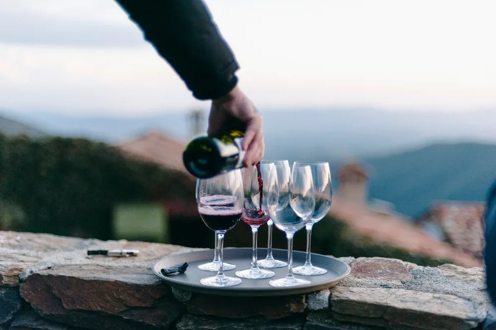 Hand pouring red wine into glasses on a tray in an outdoor setting with hills in background