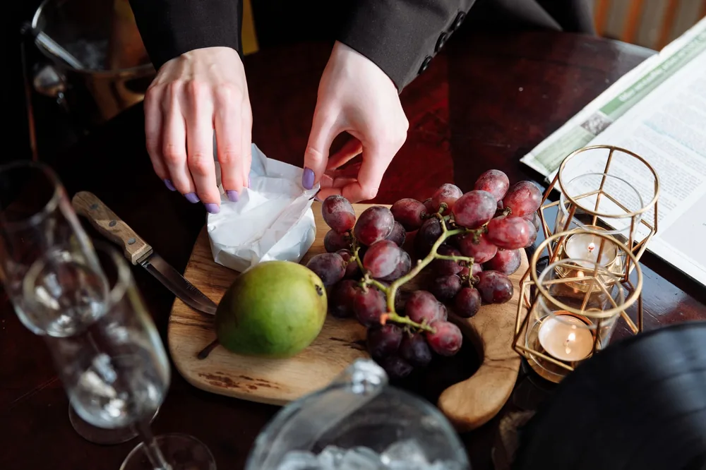 Hands arranging cheese with red grapes and a pear on a wooden board, creating an inviting spread