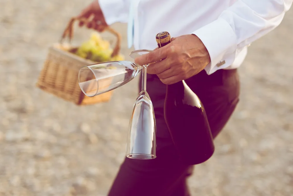 Man in white shirt carrying wine bottle, glasses, and picnic basket outdoors