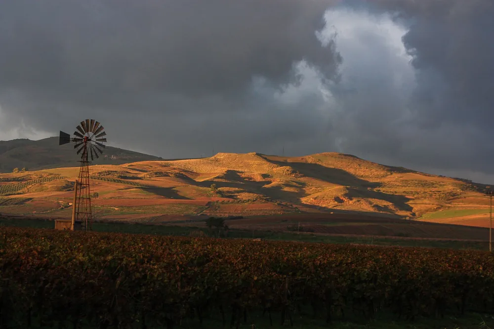 Old windmill on farm field against hill in evening sunlight and dramatic stormy sky